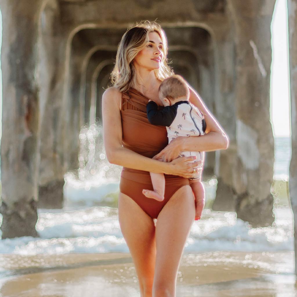 Mom holding baby by the ocean wearing Miraclesuit Network Jena One Piece Swimsuit